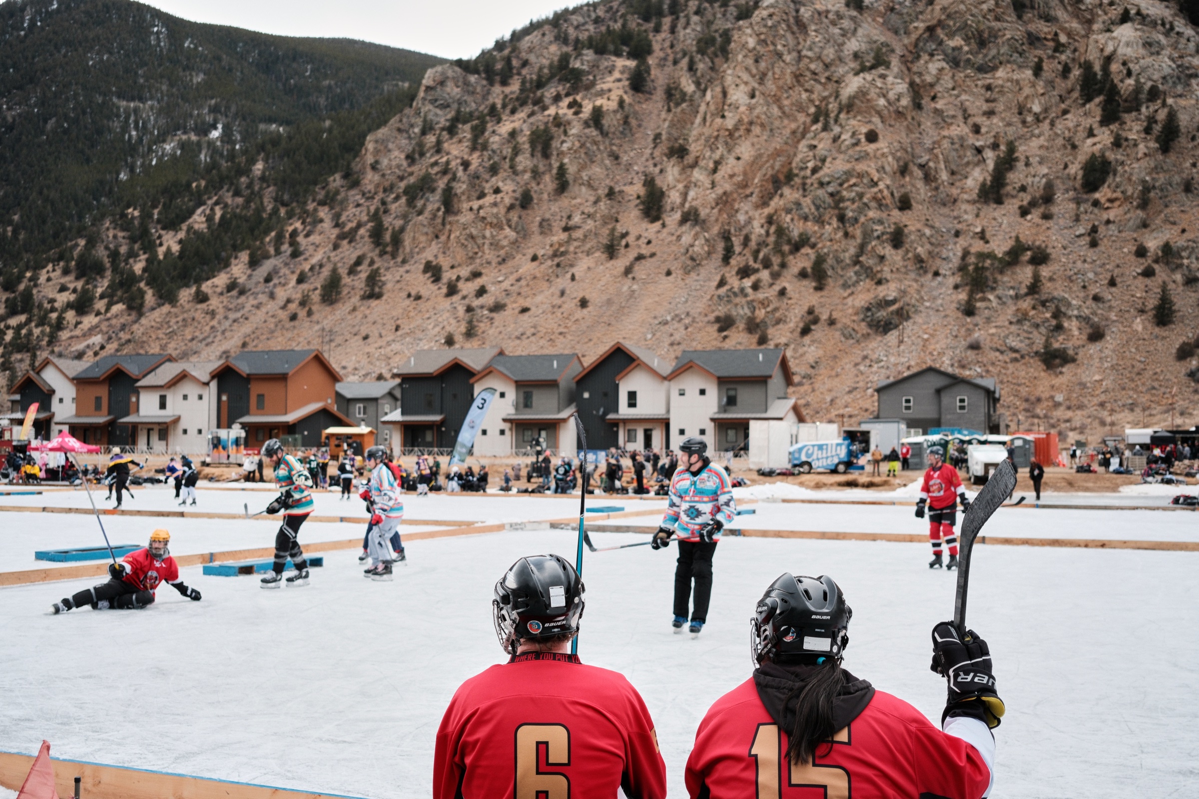 Hockey players in red watch an outdoor game on an ice rink below a steep mountain cliff