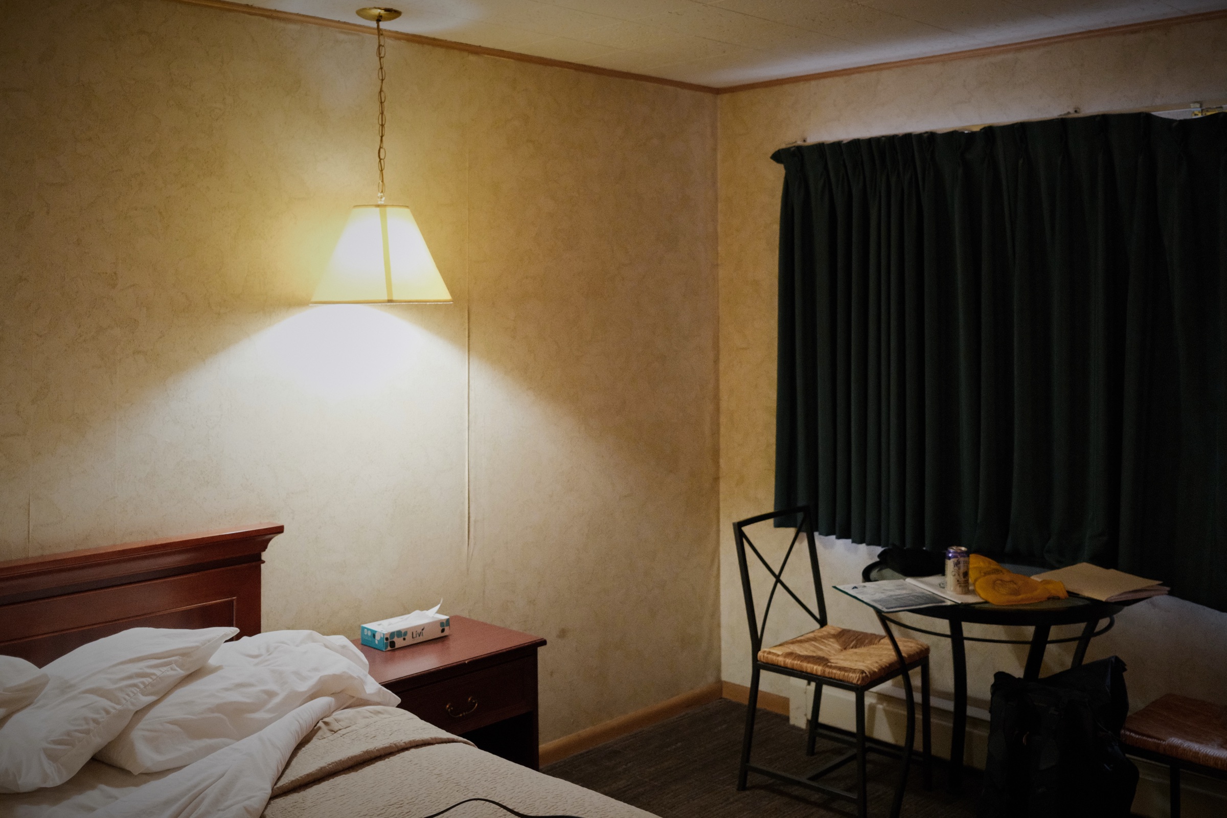 A glowing pendant lamp lights an unmade bed in a dim, dated motel room with drawn curtains and a single chair.