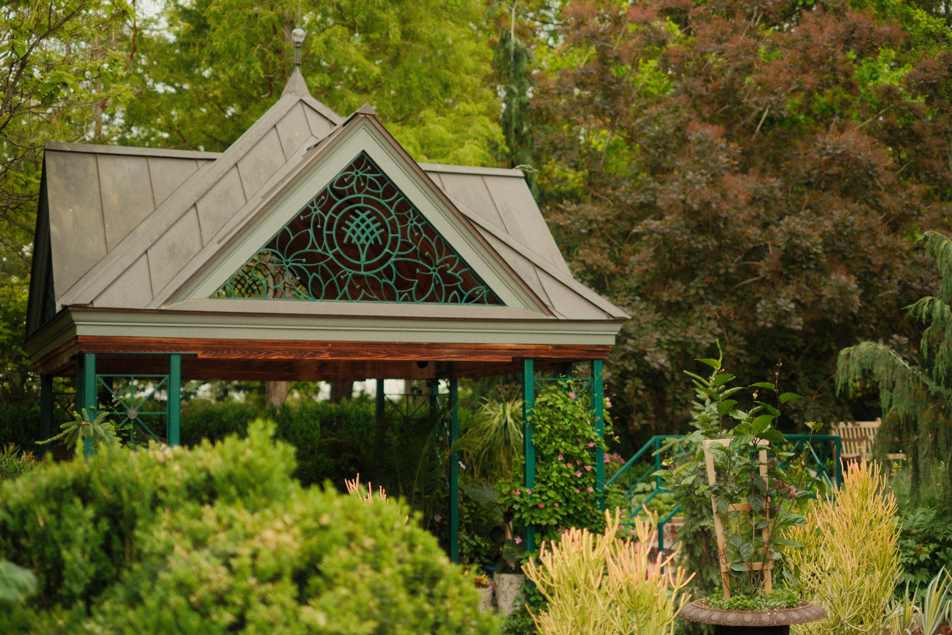 An ornate garden pavilion with a metal roof and a triangular gable featuring an intricate turquoise scrollwork design over a wooden backing. The structure is supported by green posts and surrounded by dense, manicured greenery, flowering vines, and a potted plant in the foreground.