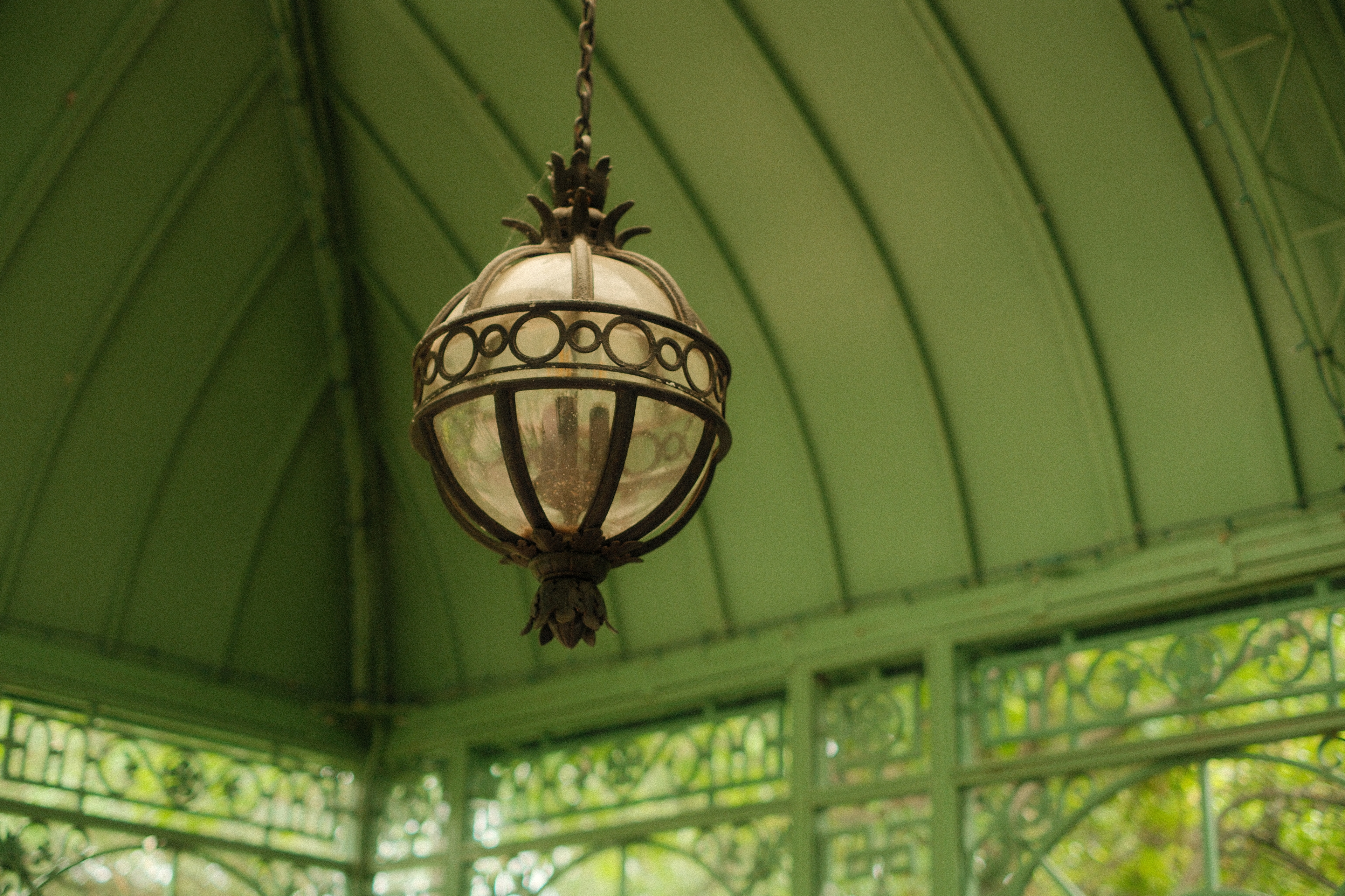 A spherical glass-and-metal pendant lantern hanging by a chain from the vaulted green ceiling of a wrought-iron conservatory. The lamp has a decorative crown-like top and a leaf-shaped finial below. Intricate ironwork windows are visible in the soft-focus background.